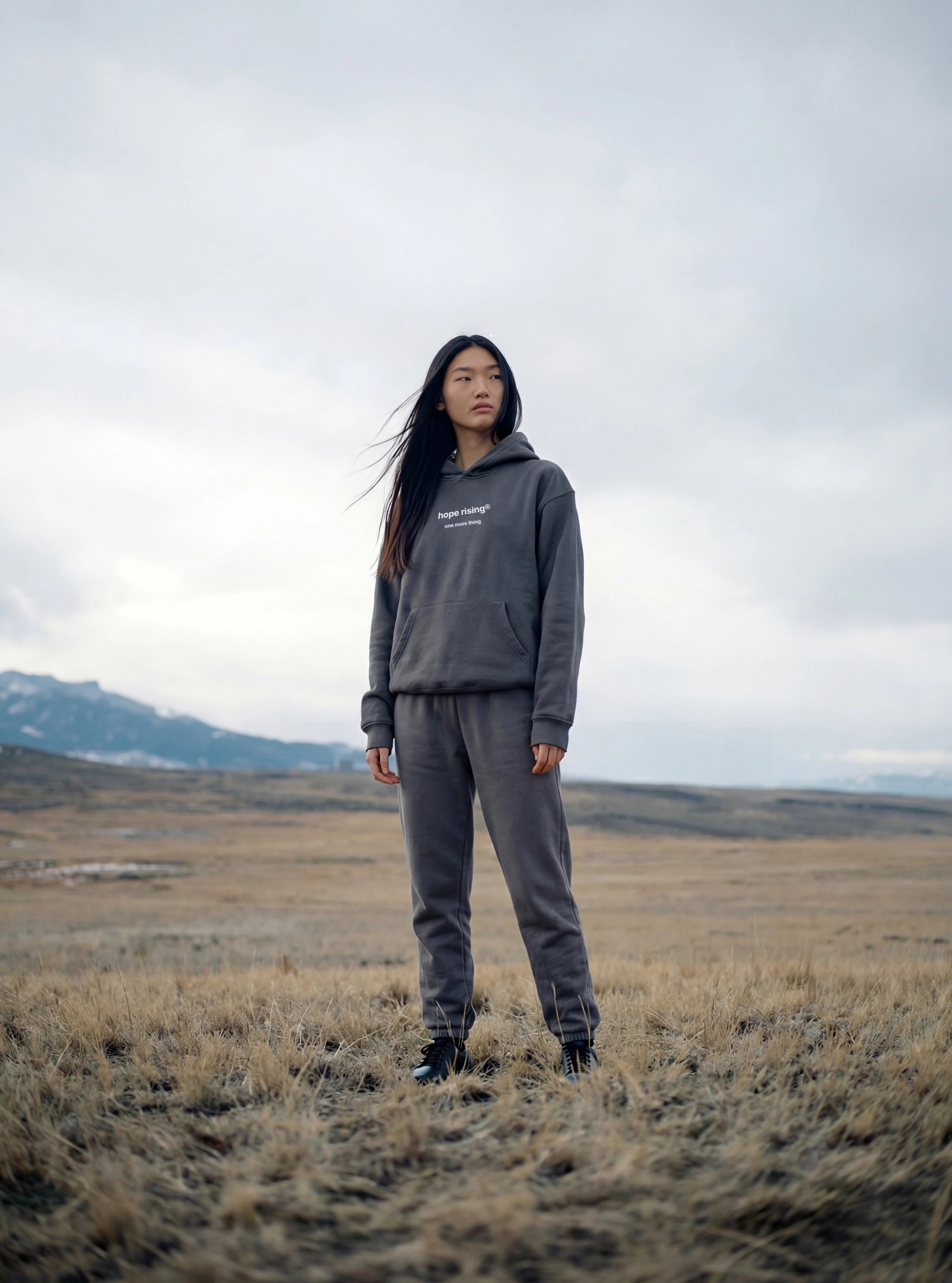 Person wearing a gray hoodie and pants standing in a field with mountains in the background