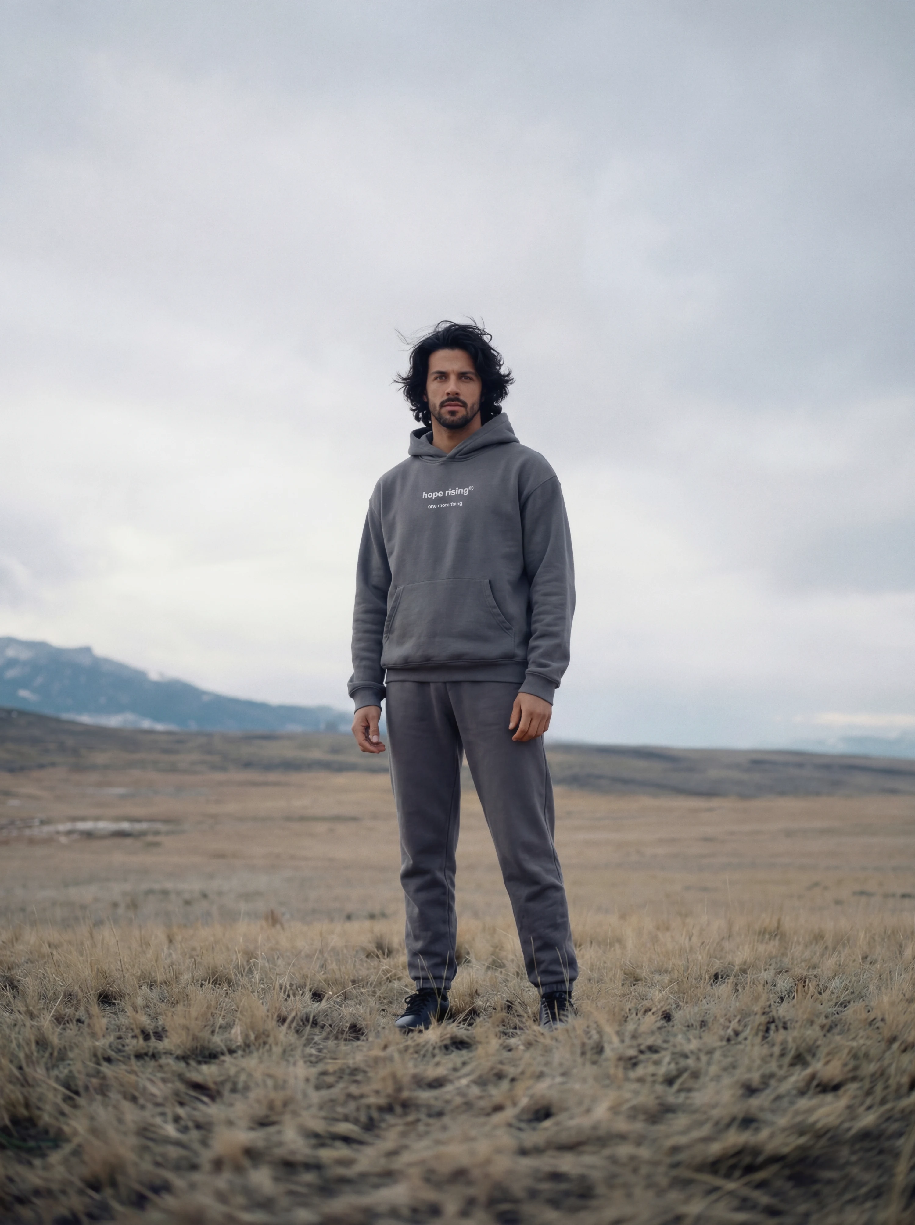Man wearing a gray hoodie and pants standing in a field with mountains in the background